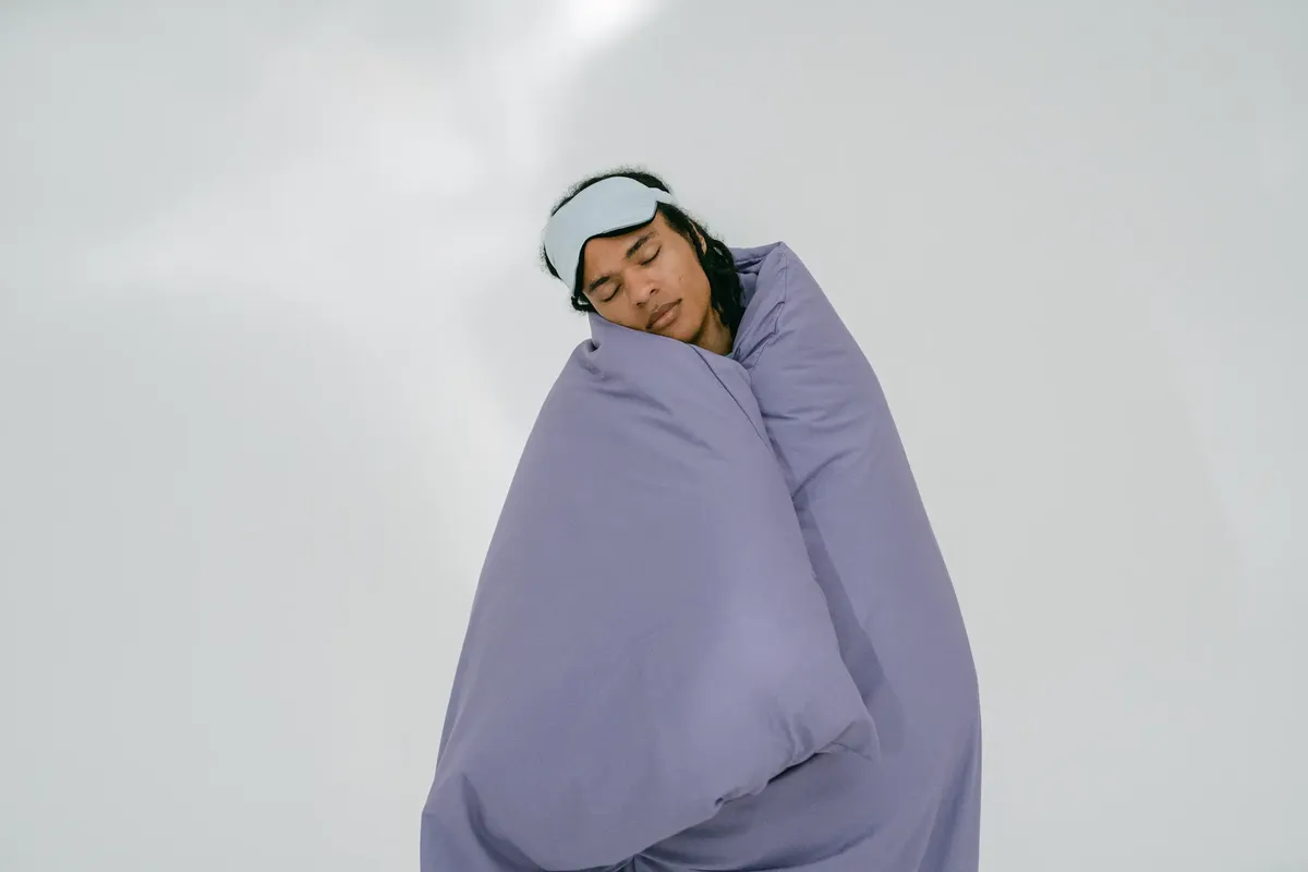 안정된 사람께서 눈 가림대를 사용하며, 흰 배경 위에 자색 뒷마스크를 덮고 침대에서 수면을 취하고 있습니다.