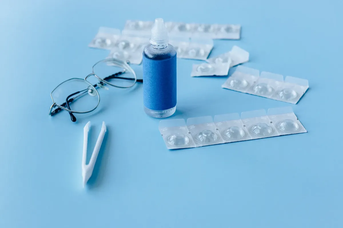 Close-up of contact lens solution bottle with eyeglasses and tweezers on blue backdrop.