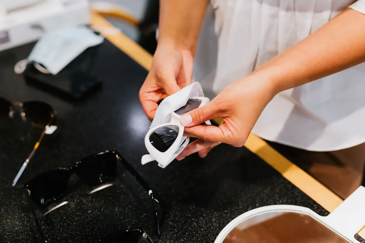 Detailed view of hands cleaning sunglasses with a lens cloth indoors on a dark surface.