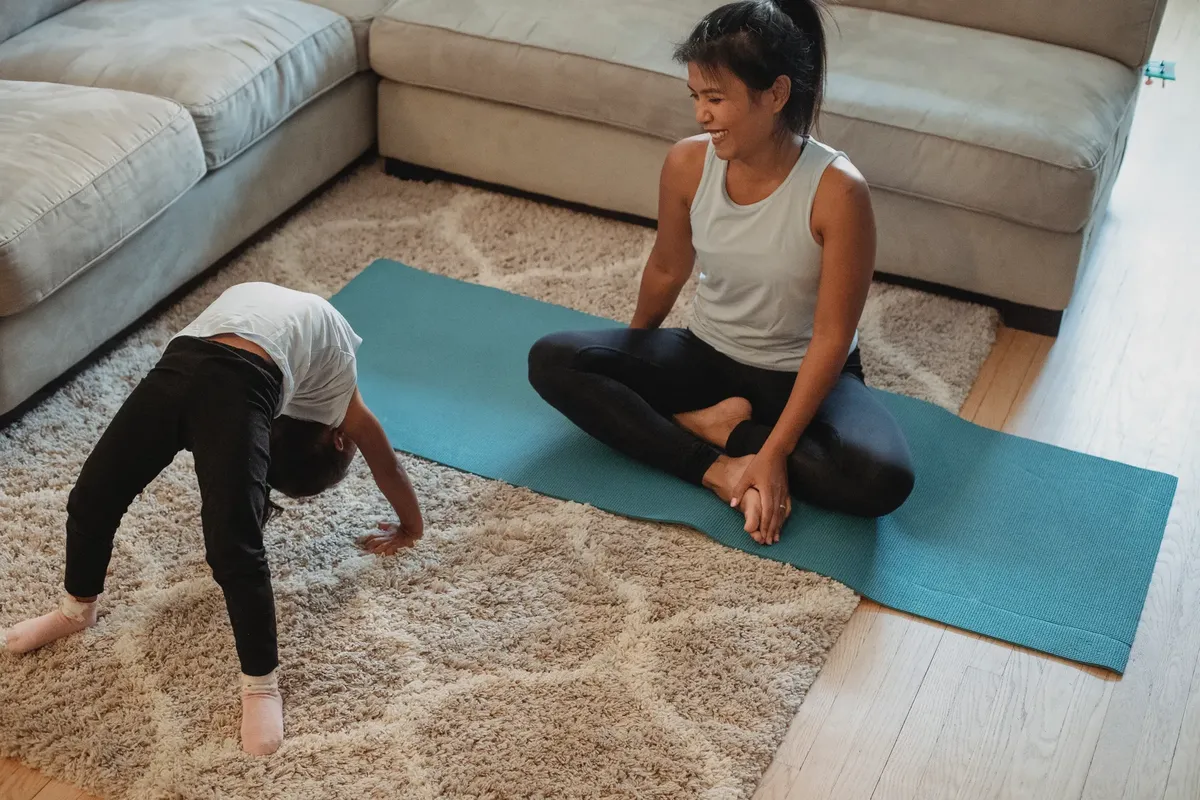 From above of happy mother sitting in lotus pose and looking at daughter bending over backwards at home in daytime