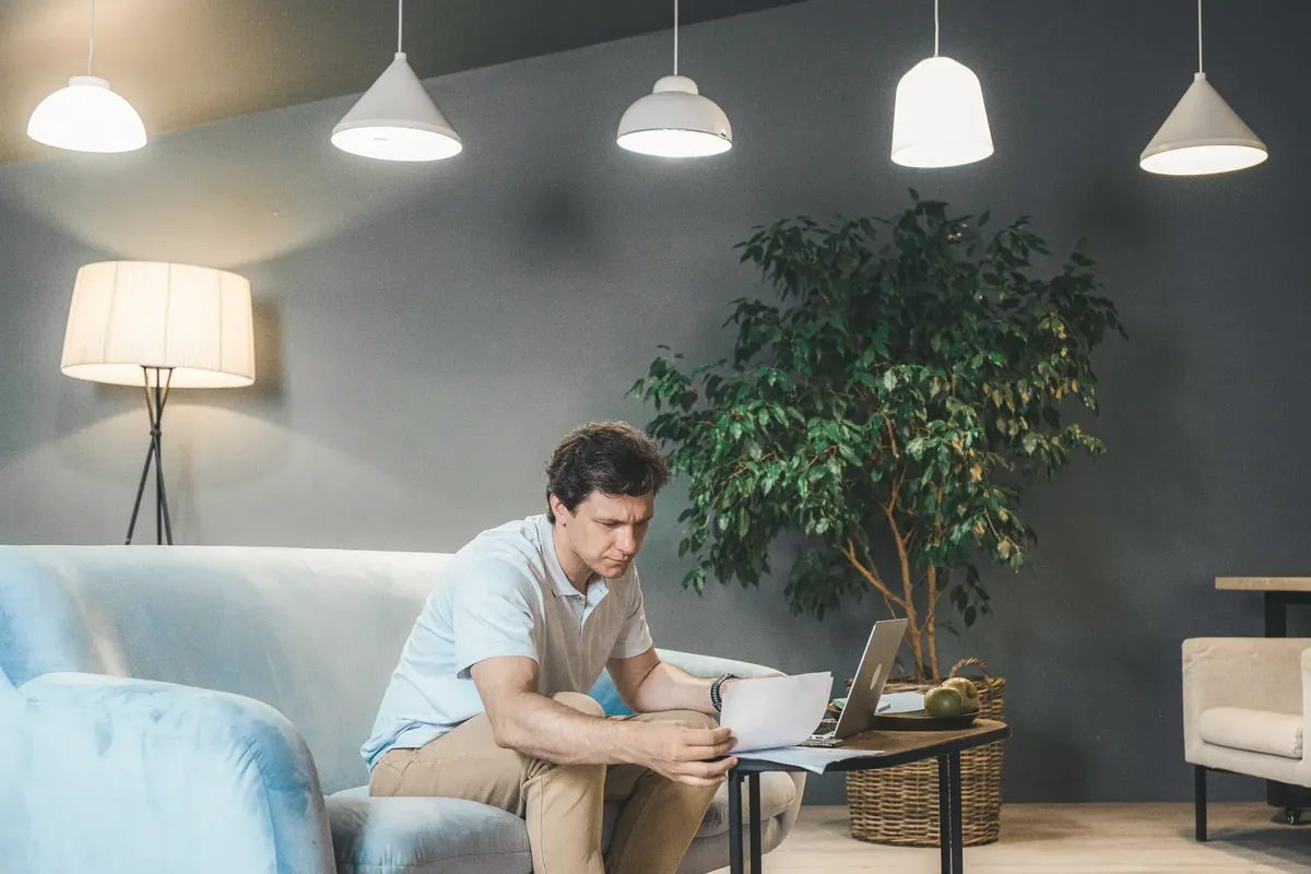 Man working on a laptop in a cozy living room with indoor plant and soft lighting.