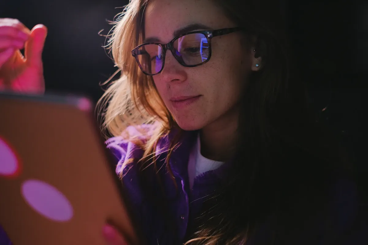 Concentrated female in eyeglasses wearing casual clothes browsing internet on modern tablet in room with low light during online work